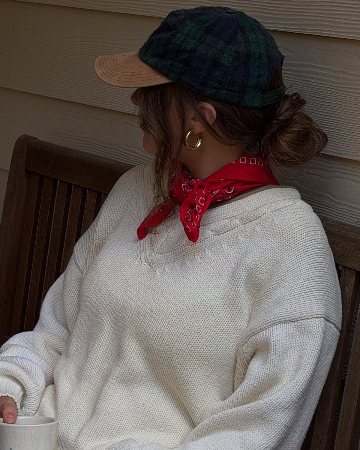 Cream cable knit sweater with a red bandana at the neck and a plaid wool cap, worn outside with a coffee.