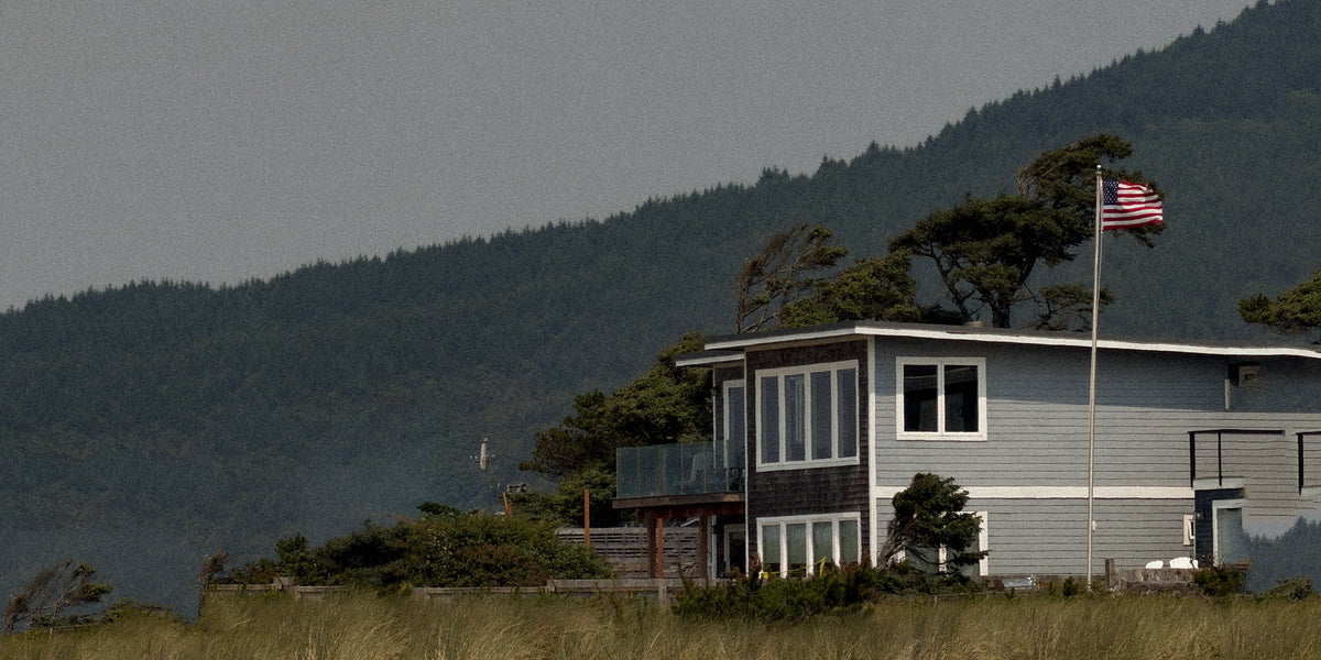 A grey shingled house on a coastal bluff with a flag flying and a forested hillside behind it.
