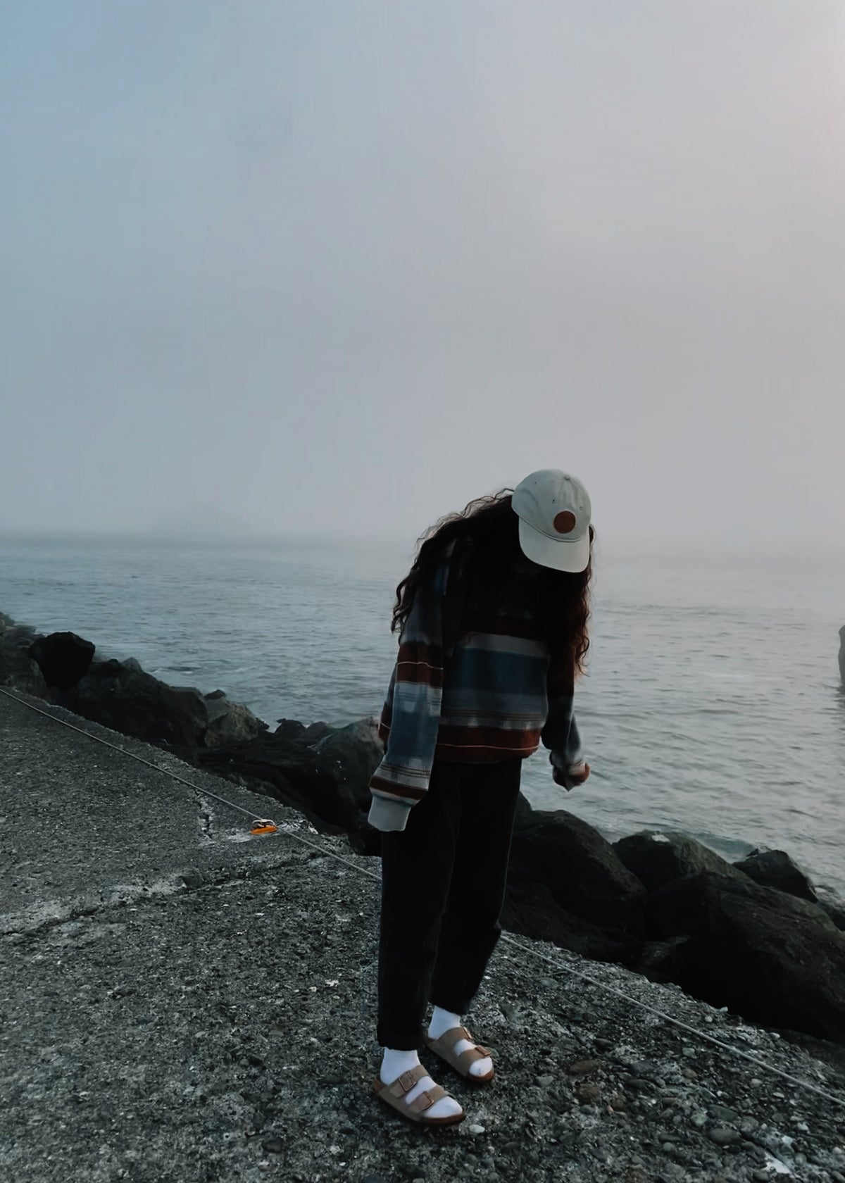 Foggy rocky shoreline with a person in a plaid flannel, ball cap and Birkenstocks, granola aesthetic outdoors