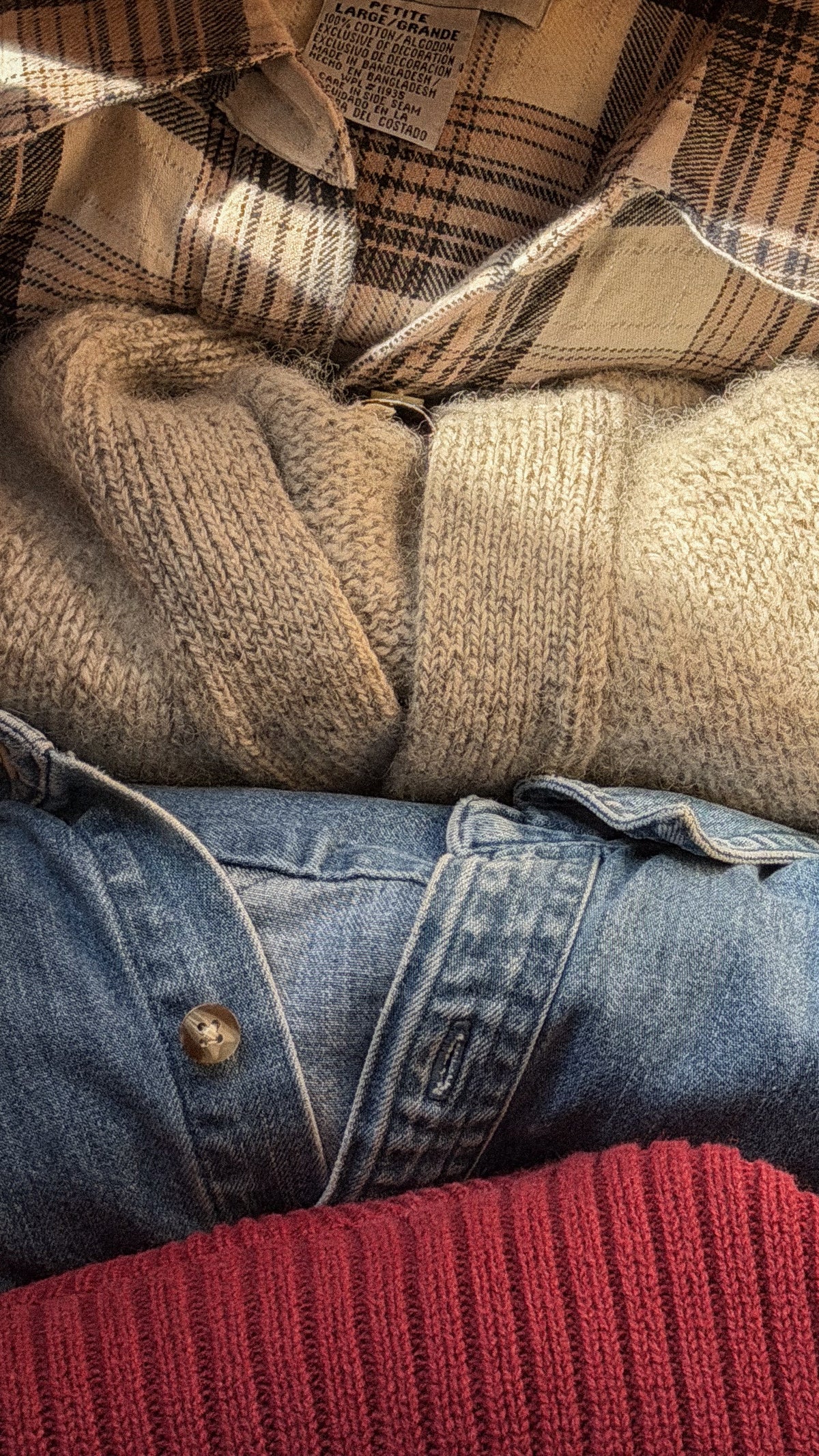 Close up stack of texture: oatmeal chunky knit, denim jacket, brown plaid flannel, red ribbed knit.