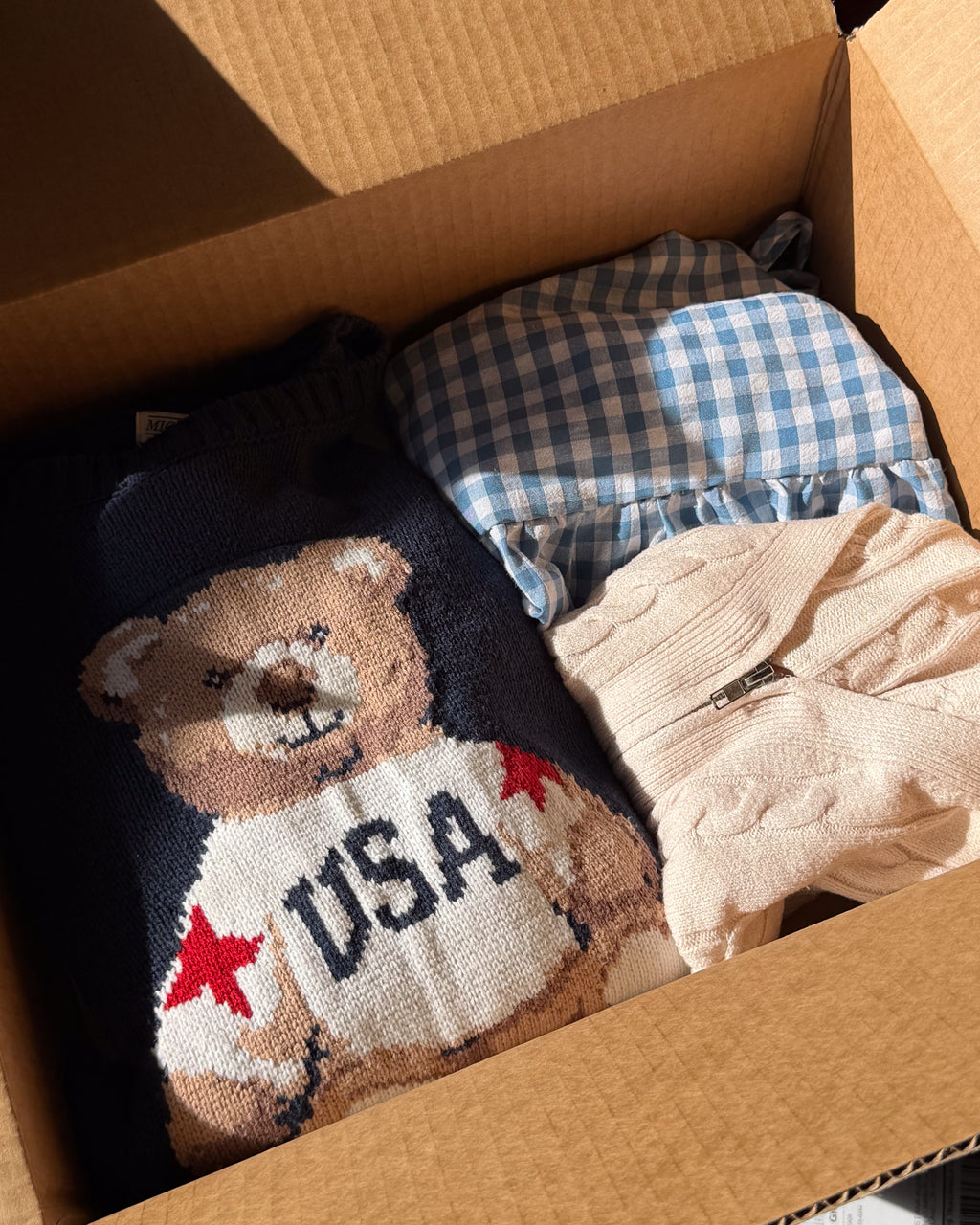 Packed box with a cream novelty bear knit, blue gingham cotton and a cream cable knit folded beside it.