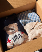 Packed box with a cream novelty bear knit, blue gingham cotton and a cream cable knit folded beside it.
