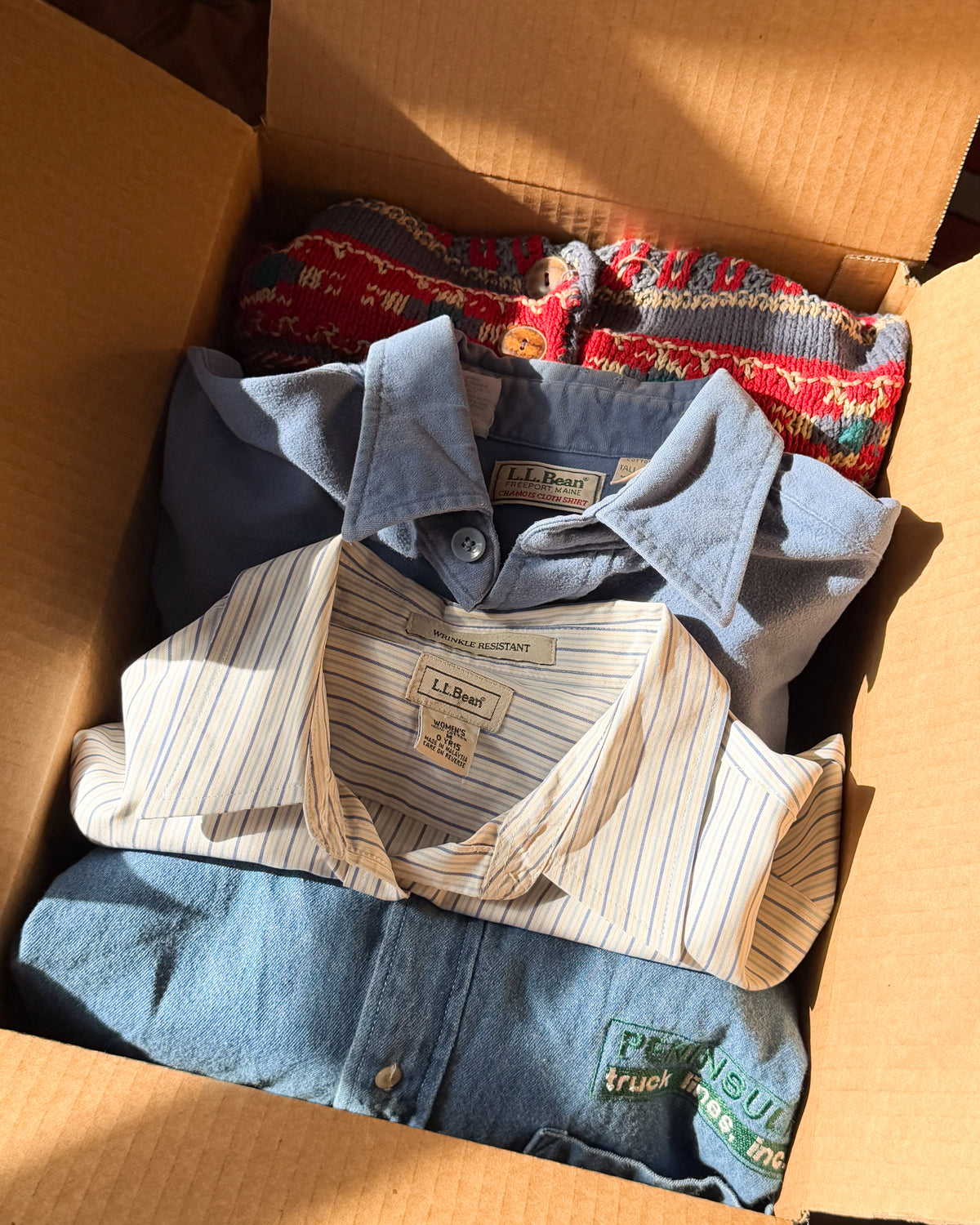 Packed box of layers: chambray shirt, cream pinstripe oxford, denim, and a red floral print folded underneath.