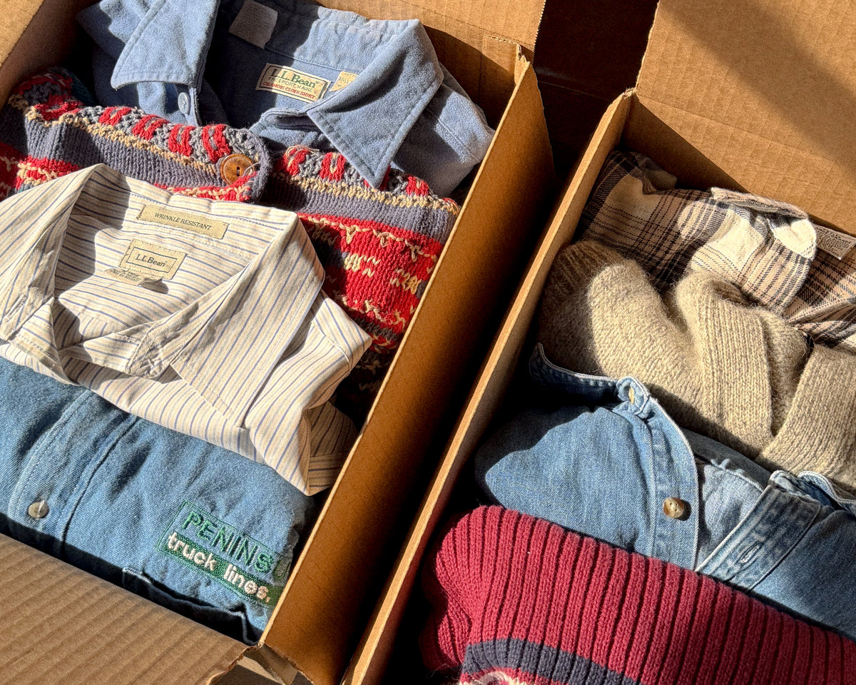 Open box of knits and shirting: pinstripe and chambray button downs, cream cable knit, denim, red ribbed sweater.