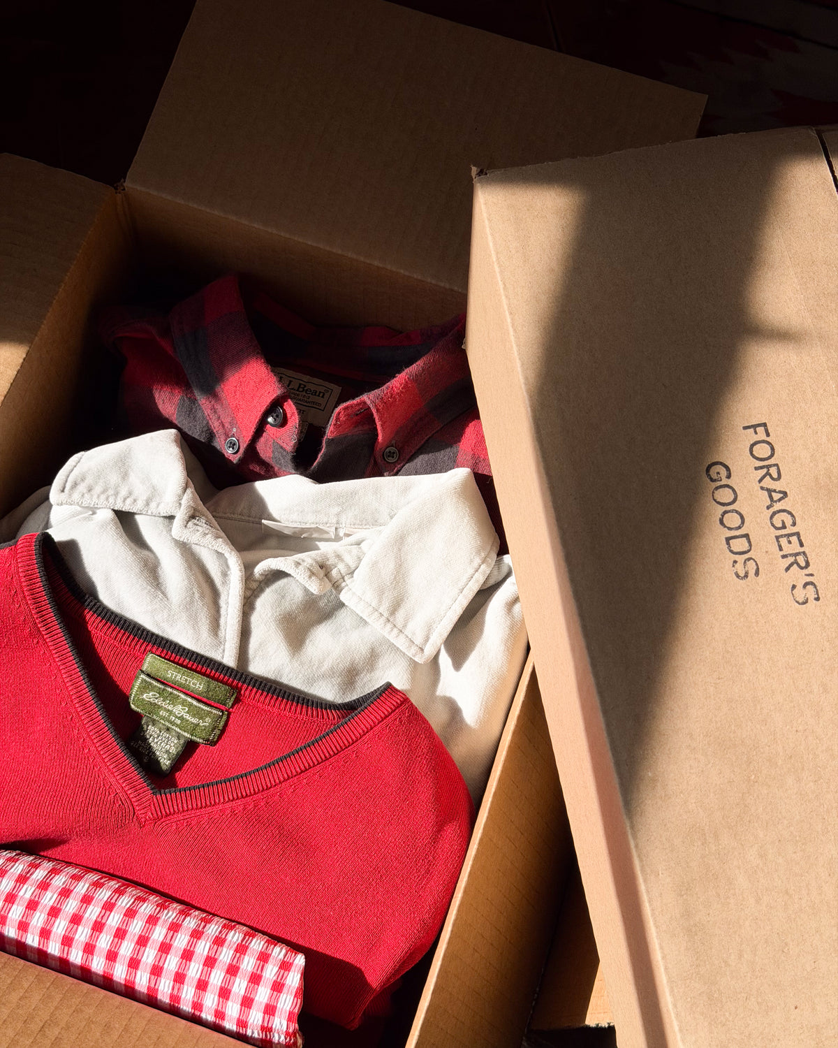 Pieces folded into a Forager's Goods box: red v neck sweater, cream collared shirt, red gingham cotton.