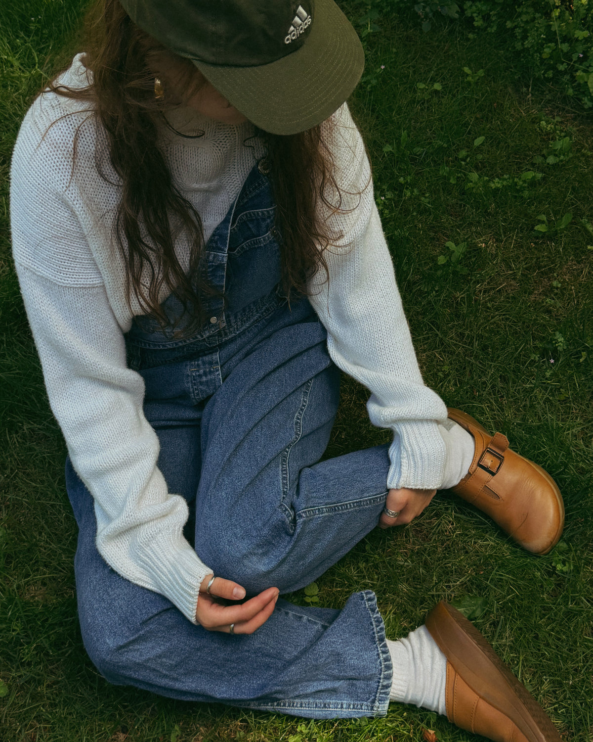 Sitting on the grass in denim overalls, a white knit sweater, a green cap and tan clogs.